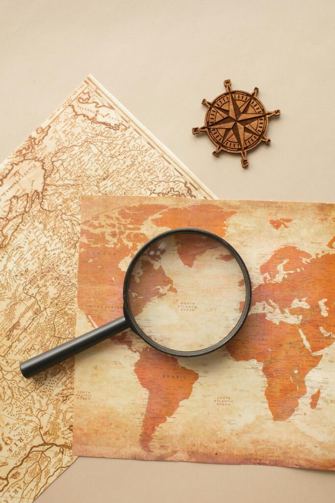 Top view of round magnifying glass on paper maps placed on beige background with wooden wind rose in light room