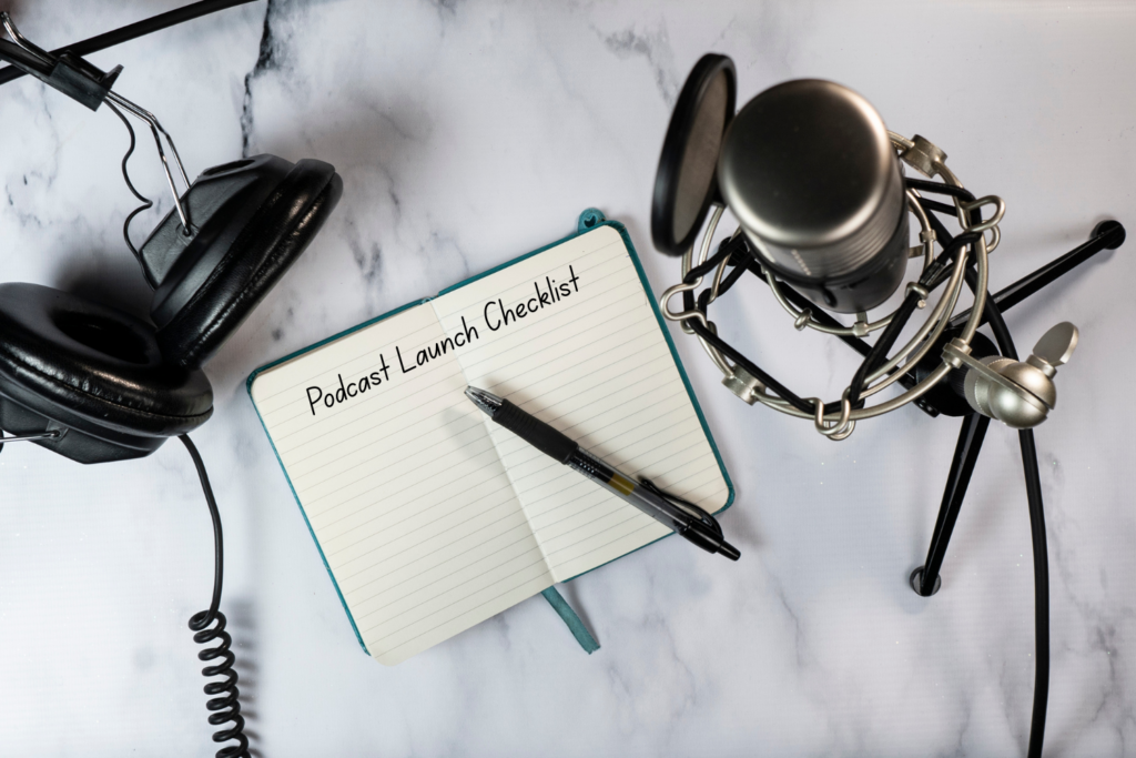 Podcast launch checklist notebook beside a microphone and headphones on a white marble desk.