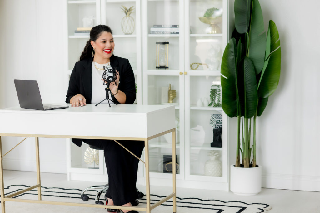 Latina woman sitting at a white desk with her hand over the microphone, looking to the right. 