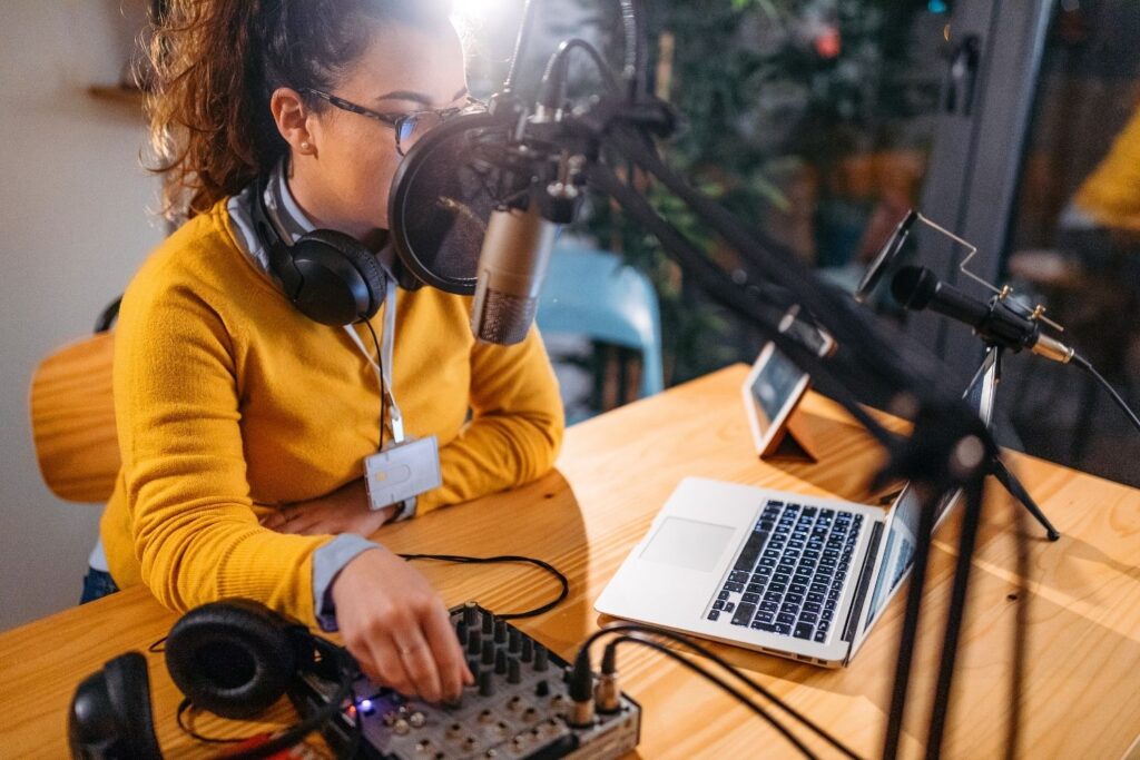 Podcast Ideas: How to Find Yours and Build a Show Worth Launching A medium close-up photograph of a woman with curly hair and glasses, wearing a yellow sweater, actively producing a podcast at a wooden desk. She speaks into a microphone with a pop filter and adjusts a knob on an audio mixer, with a laptop, headphones, and another microphone also on the desk in an indoor setting.