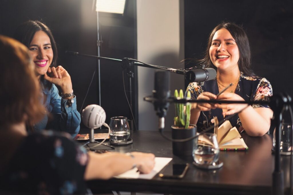 A candid medium shot of two diverse women smiling and laughing together at a professional studio desk with multiple dynamic microphones, recording a podcast. Another woman's back is visible in the foreground. Water glasses, notebooks, and dynamic microphones populate the table against a dark, well-lit background.