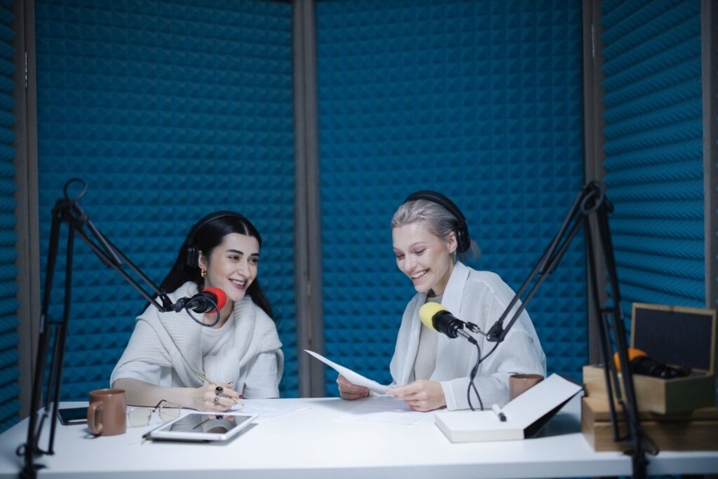 two women wearing headphones and speaking into professional microphones record a podcast episode in a studio with blue acoustic foam paneling. The woman on the right holds a script and smiles while the woman on the left looks down at a tablet.