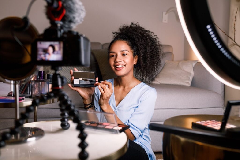 A candid medium shot of a smiling Black woman with prominent curly hair tied up, wearing a light blue shirt, filming a beauty vlog tutorial. She holds an eyeshadow palette and brush, looking at an off-camera setup. A professional DSLR camera on a flexible tripod with a shotgun microphone is visible in the foreground to the left. A large LED ring light is on the right, and other makeup products are on the table against a grey couch.