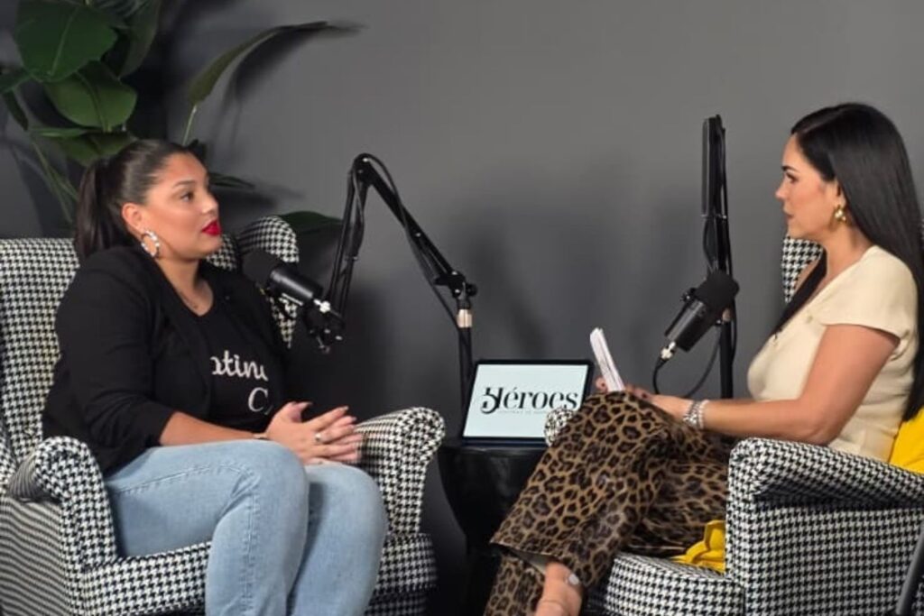 A photograph of two women recorded for a podcast interview in a modern studio. The image features Scarlet Ortiz and Heydi de la Cruz, a podcast coach and podcast editor, seated in matched houndstooth armchairs. One woman, in a black shirt and jeans, is speaking into her microphone, while the other, in a beige top and leopard-print pants, holds papers. A small table with equipment and a tablet showing "Héroes" is between them, set against a grey wall with a large potted bird of paradise plant.